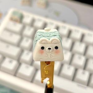 May include: A white and green keycap with a cute cartoon animal face. The keycap is attached to a gold-colored keycap puller.