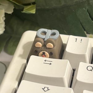 May include: A close-up of a white mechanical keyboard with a custom keycap featuring a bear design. The bear keycap is brown with a blue bow and a small red tongue. A yellow knitted item with white accents is in the background.