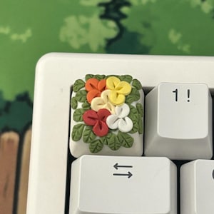 May include: A white keyboard with a keycap featuring a small, colorful flower arrangement made of polymer clay. The flowers are red, orange, yellow, and white. The keycap is on the left side of the keyboard, above the arrow keys.