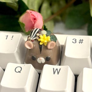 May include: Close-up of four custom keycaps with mouse designs. The keycaps are beige, brown, and black, each with a mouse face and a floral crown. They are on a white keyboard, with pink roses and green leaves in the background.