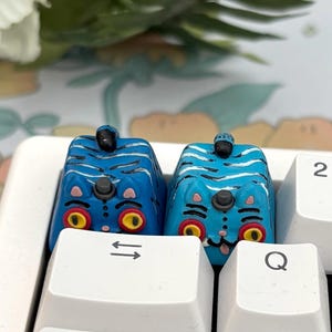 May include: Two blue and white artisan keycaps with a tiger design are installed on a white keyboard. The keycaps have yellow eyes and black stripes. The keyboard keys include the symbols for the up and down arrows, the letter Q, the letter A, and the number 2.