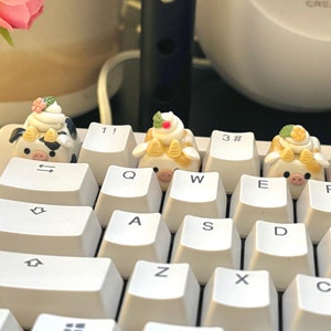 May include: A white keyboard with custom keycaps shaped like cartoon cows with desserts on their heads. The keycaps are on the Q, W, E, and 1 keys. A pink flower is in the background.