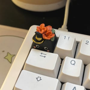 May include: A black artisan keycap with a cat face, a witch hat, and orange flowers. The keycap is on a white keyboard, next to the number 1 key. The cat has a cute expression.