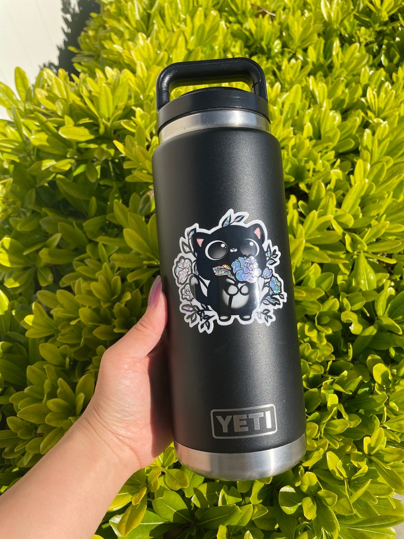 May include: A black Yeti water bottle with a sticker of a black cat holding flowers. The sticker has a holographic finish.