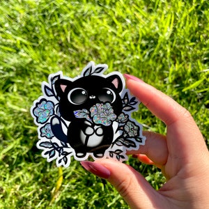 May include: A black and white sticker of a cartoon cat holding a bouquet of iridescent flowers. The cat has large eyes and a small mouth. The flowers are surrounded by leaves and vines.