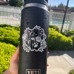 May include: Black Yeti water bottle with a black and white sticker of a cartoon cat holding flowers. The sticker has a glittery finish.