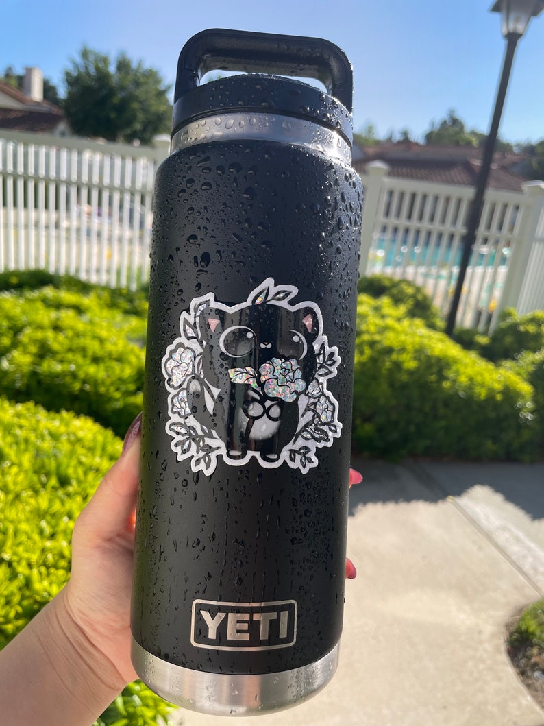 May include: Black Yeti water bottle with a black and white sticker of a cartoon cat holding flowers. The sticker has a glittery finish.