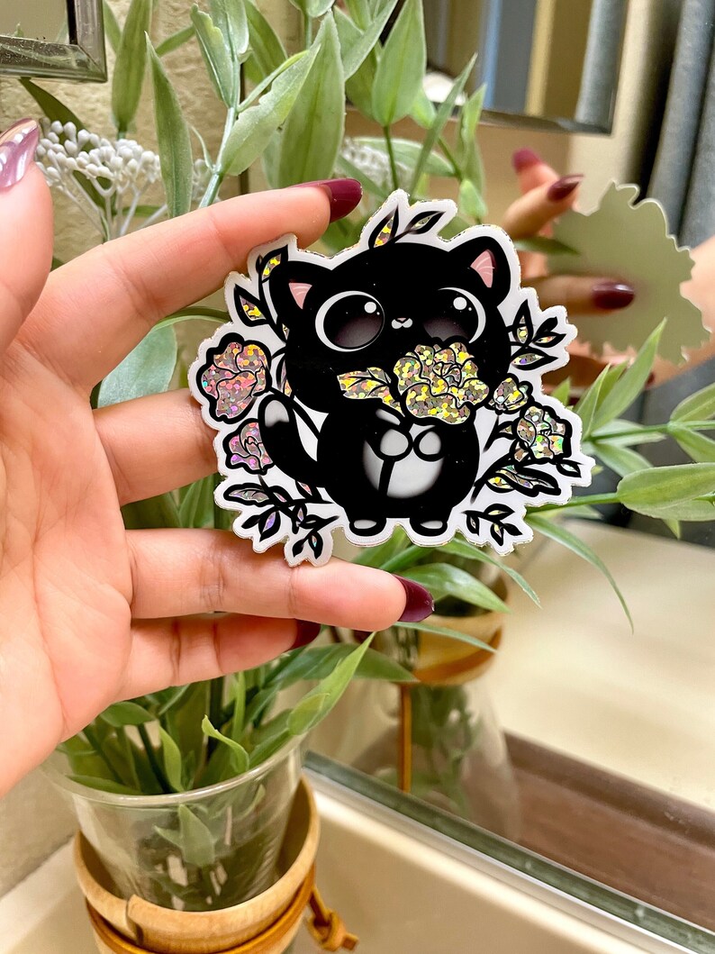 May include: A black cat sticker with glitter accents, holding a bouquet of pink and yellow flowers. The sticker is on a white background.