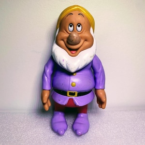 Può includere: Una statuetta di un nano vestito di viola con una barba gialla e un viso marrone. Il nano ha un ampio sorriso e grandi occhi marroni.