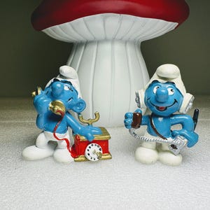 May include: Two blue Smurf figurines, one with a red and gold telephone, the other with headphones and a walkie-talkie. A large mushroom-shaped decoration with a red top and white stem is in the background.