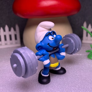 May include: A blue Smurf figurine lifting a gray barbell. The Smurf is wearing a white hat, black shoes, and a brown vest with a yellow belt. A large mushroom and a white picket fence are in the background.