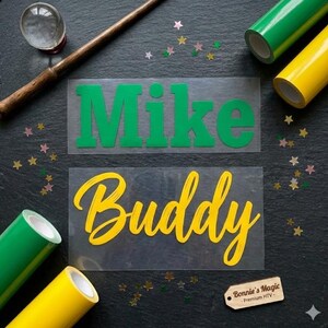 May include: Two heat transfer vinyl (HTV) designs with the names "Mike" in green and "Buddy" in yellow. Rolls of green and yellow HTV are visible, along with a wooden wand, a crystal ball, and star-shaped confetti. A small tag reads "Bonnie's Magic - Premium HTV".
