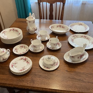 May include: A collection of vintage white porcelain dishware with a pink floral pattern. The set includes plates, bowls, teacups, saucers, a teapot, a sugar bowl, and a gravy boat, all arranged on a wooden table.