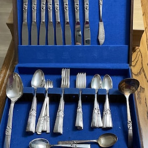 May include: A vintage silverware set displayed in a wooden case with a blue velvet lining. The set includes knives, forks, spoons, and a ladle, all with ornate silver handles. The case is open, revealing the neatly arranged utensils.