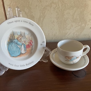 May include: A white ceramic plate and teacup set featuring illustrations of rabbits. The plate reads "Once upon a time there were four little Rabbits." The teacup and saucer also have rabbit illustrations.