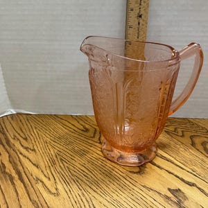May include: A vintage pink glass pitcher with a floral pattern. The pitcher has a handle and a spout for pouring. It is sitting on a wooden surface. A wooden ruler is behind the pitcher.
