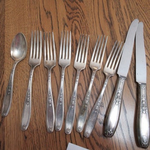 May include: A set of silverware, including a spoon, forks, and knives, all with ornate handles. The silverware is made of silver and has a polished finish.