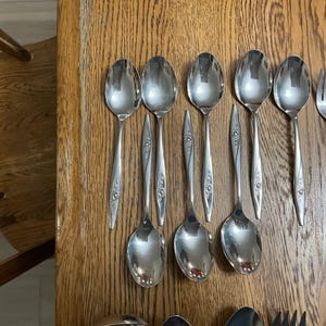 May include: A collection of vintage silverware, including spoons, forks, and knives, arranged on a wooden surface. The silverware has a floral design on the handles and a shiny, reflective finish. The spoons are oval-shaped.