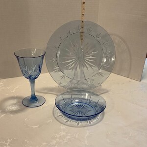 May include: A light blue glass dinnerware set, including a wine glass, a dinner plate, and a small bowl. The plate and bowl have a starburst design. The wine glass is approximately 7 inches tall.