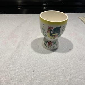 May include: A white ceramic egg cup with a yellow rim and a decorative rooster and floral design. The egg cup has a pedestal base and is likely vintage. The design includes red, green, and blue accents.
