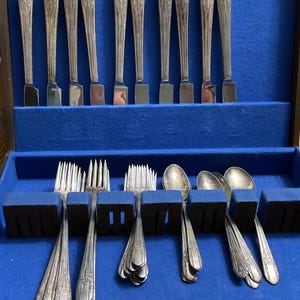 May include: A set of vintage silverware, including knives, forks, and spoons, arranged in a blue velvet-lined case. The silverware has ornate floral detailing on the handles. The case is open, revealing the neatly organized utensils, perfect for a formal dining setting.
