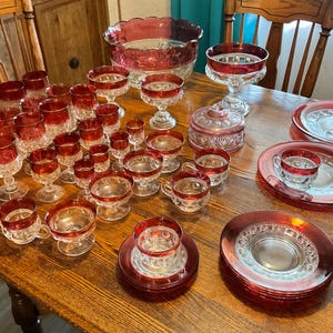 May include: A collection of ruby red and clear glass tableware, including glasses, cups, plates, and serving bowls, arranged on a wooden table. The glassware features a decorative rim and base. The set includes various sizes, suitable for a formal dining setting.