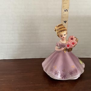 May include: A porcelain figurine of a woman in a lavender dress holding a bouquet of pink flowers. The dress is adorned with floral accents. The figurine is approximately 4.5 inches tall.