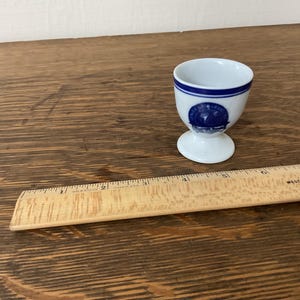 May include: A white ceramic egg cup with a blue band and a blue and white design. The egg cup has a pedestal base.