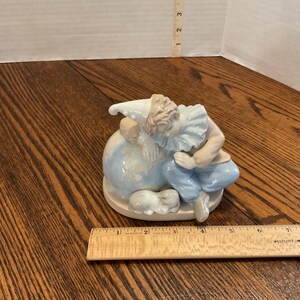 以下が含まれることがあります： 淡い青色の地球儀の上に休んでいるピエロの磁器製置物。ピエロは白い帽子、フリルカラー、青いズボンを着用しています。小さな白い動物が地球儀の底にいます。置物はクリーム色の台座にあります。