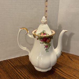 May include: White porcelain teapot with a floral design featuring red, pink, and yellow roses. The teapot has a gold-colored handle and trim, and a matching lid with a finial. A vintage tea set item.