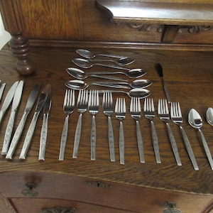 May include: A set of silverware laid out on a wooden surface. The set includes knives, forks, and spoons. The silverware is made of stainless steel and has a polished finish.