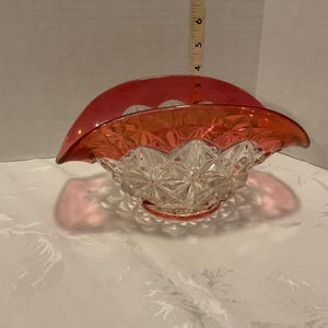 May include: A vintage, clear glass basket with a ruby red rim and handle. The basket has a starburst pattern on the bowl and a scalloped edge. The handle curves over the top. A decorative piece.