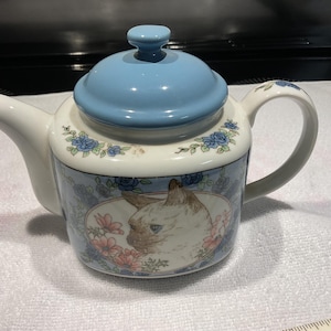 May include: A white ceramic teapot with a light blue lid and handle. The teapot features a decorative design with a cat portrait, blue flowers, and pink flowers. The spout and handle are white.