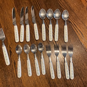 May include: A collection of vintage silverware, including knives, spoons, and forks. The utensils have white handles with a delicate floral pattern in blue and green. The stainless steel blades and tines gleam against the wooden surface.