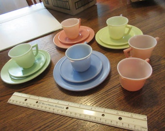 May include: A collection of pastel-colored glass tea cups and matching saucers in shades of pink, blue, and green. A white plastic ruler with Bell & Howell Schools branding is positioned in the foreground, showing a length of 12 inches for scale.