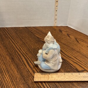 以下が含まれることがあります： 白い帽子と水色とベージュの衣装を着た、座っているピエロの磁器製置物。ピエロは小さなフィギュアを持っています。置物は白い台座の上にあります。