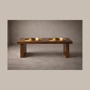 May include: A wooden pet feeder with two gold-colored bowls. The feeder is made of dark brown wood and has a rectangular shape with two circular cutouts to hold the bowls. The bowls are made of a shiny gold metal. The feeder is elevated on two rectangular legs.