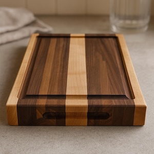 May include: A rectangular wooden chopping board with a light-coloured border and a central panel of alternating dark and light wood strips. The board has a juice groove and finger grips. The wood grain is visible.