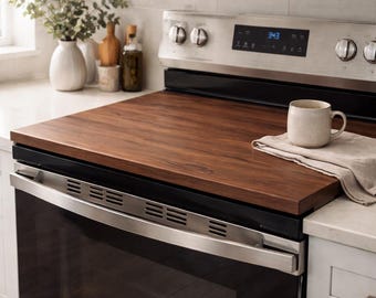 May include: A dark brown wooden stove top cover sits atop a stainless steel oven. A beige mug rests on a linen cloth. The kitchen setting includes a white vase with greenery, and bottles of oil and vinegar.