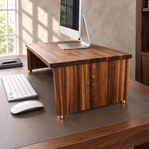 May include: A rectangular monitor stand crafted from dark brown wood with a visible grain pattern. The stand features four small, cylindrical gold-toned metal feet at the base, providing elevation for a computer monitor placed on top.