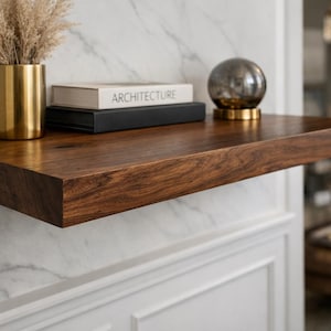 May include: A floating wooden shelf with a rich, dark brown finish. The shelf holds a gold vase with dried plants, two books, and a decorative glass sphere on a gold base. The shelf is mounted on a white and gray marble wall.