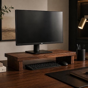 May include: A dark wood monitor stand with a black computer monitor, keyboard, and mouse on a wooden desk. A gold desk lamp and black pen holder are also on the desk. A vase with greenery and books are on the left.