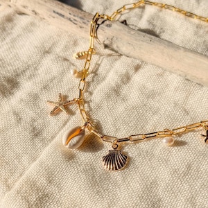 May include: A gold-colored necklace with various charms, including a starfish, seashell, and pearl accents. The chain is a link style, and the necklace is displayed on a neutral-colored fabric. The charms are detailed and add a beachy theme.