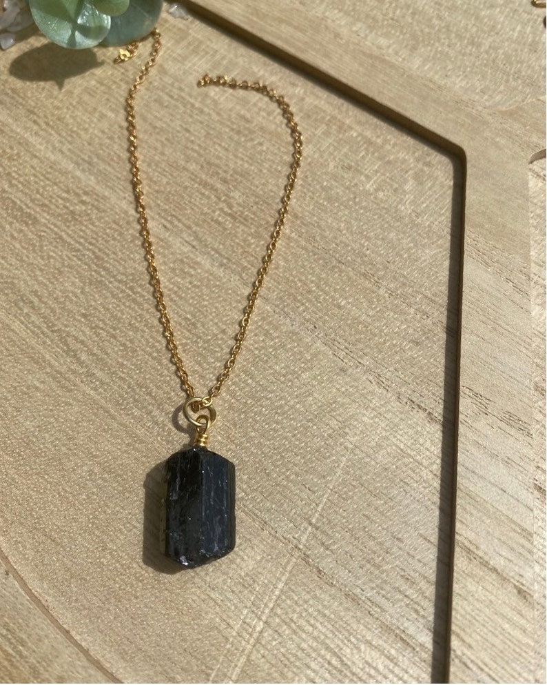 以下が含まれることがあります： 暗い長方形の黒い石のペンダントが付いた、金色のチェーンネックレス。 ネックレスは、明るい茶色の木製の表面に表示されています。 ペンダントは、ラフカットの天然石です。