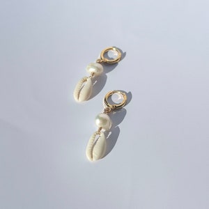 Könnte beinhalten: Ein Paar goldfarbene Creolen mit einem hängenden Design. Jeder Ohrring hat eine weiße Muschel und eine weiße Perle. Die Ohrringe werden auf einer weißen Oberfläche mit Schattenwurf präsentiert.