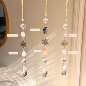 Può includere: Tre acchiappasole di cristallo sospesi con catene dorate. Ognuno presenta una varietà di pietre preziose, tra cui quarzo rosa, agata muschiata, citrino, ametista e pietra di luna. Lavorazioni metalliche decorative e pendenti di cristallo trasparente completano il design.