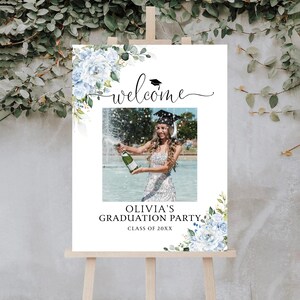 May include: A white graduation party sign on an easel, with a photo of a graduate popping champagne. The sign has floral accents, the word "welcome", and text that reads "Olivia's Graduation Party, Class of 20XX".