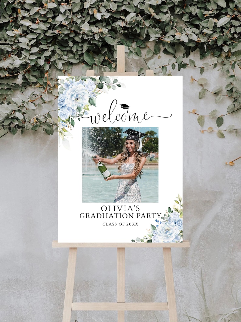 May include: A white graduation party sign on an easel, with a photo of a graduate popping champagne. The sign has floral accents, the word "welcome", and text that reads "Olivia's Graduation Party, Class of 20XX".