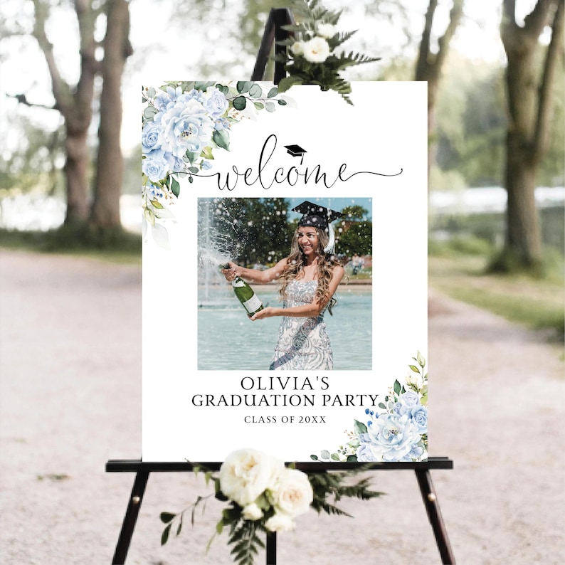 May include: A white graduation party welcome sign on a black easel. The sign features a photo of a graduate popping champagne, floral accents, and the text "Welcome" and "Olivia's Graduation Party".