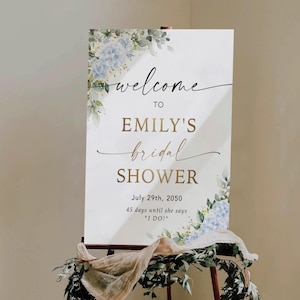 May include: A white sign with the words "welcome to Emily's bridal shower" in elegant script. The sign is decorated with blue floral accents and greenery. The date July 29th, 2050, is displayed, along with the text "45 days until she says 'I DO!'"